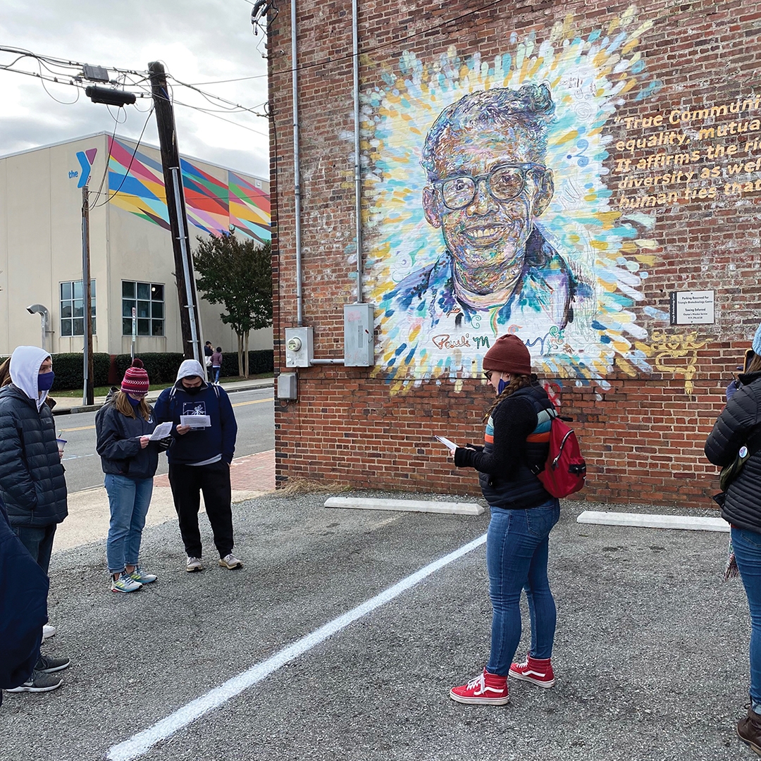 A group of five people look down, reading a paper with a large colorful mural of Rev. Dr. Pauli Murray in the background in downtown Durham