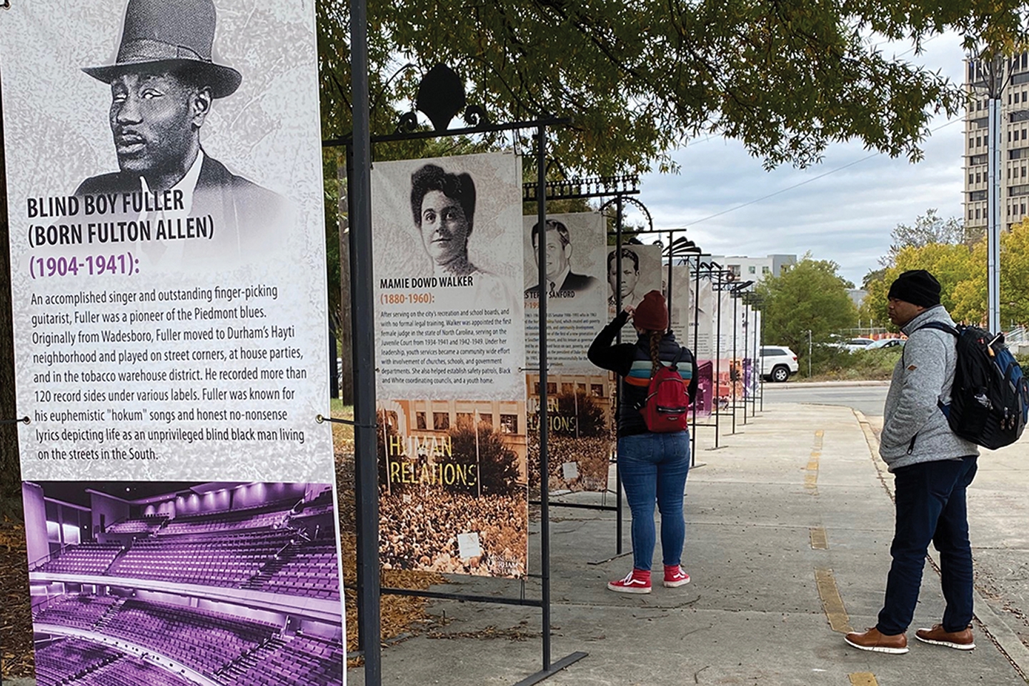 Two people read large signs featuring Blind Boy Fuller and Mamie Jo Walker in downtown Durham