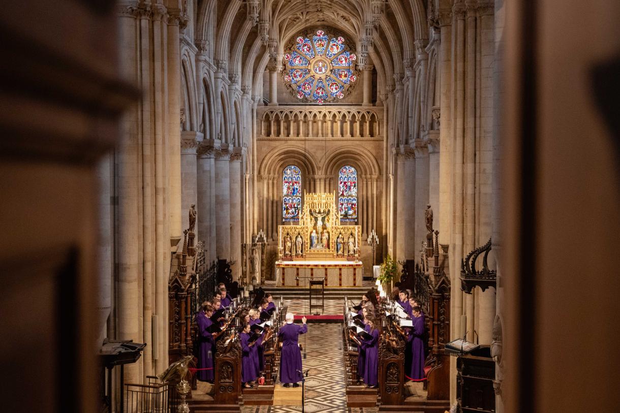 A high-angle, wide shot looking down the main aisle of a large, gothic-style church during a choir performance. The choir members, dressed in purple robes, are arranged in two sections of choir stalls facing each other in the midground. A conductor in a purple robe stands on a raised platform between them.