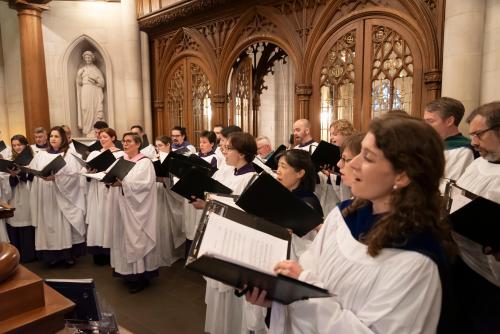 A large choir of about twenty people, dressed in white robes with some wearing dark blue or purple stoles, stands in a church and sings from black binders of sheet music.