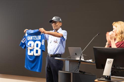 A medium shot captures an older man standing on a stage, holding up a bright blue sports jersey with the name "DANTZLER" and the number "28" printed in white on the back. He wears a light blue button-down work shirt, dark pants, a baseball cap, and an ID badge. To his right, a woman with blonde hair stands behind a podium equipped with a laptop and monitor, smiling and applauding the man.