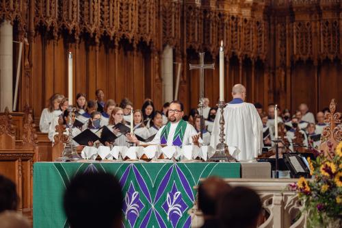 A medium shot of a religious service in a cathedral. A clergyman in a white robe and green stole stands behind a large altar covered with a green and purple patterned cloth. He is leading a prayer with his hands outstretched.