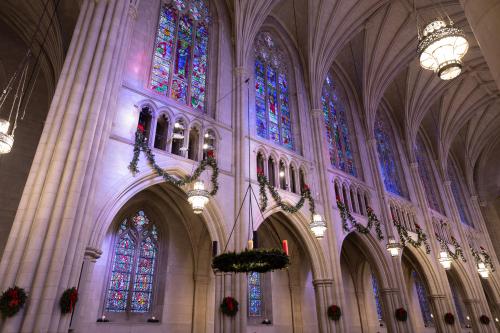  A low-angle, interior shot captures the architecture of a Gothic-style chapel nave, decorated for the Christmas season. The image focuses on the tall stone walls, which feature multiple levels of pointed arches and lancet stained-glass windows, bathing the space in colorful light. 