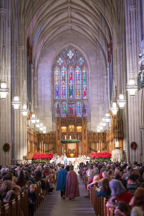  A vertical, wide-angle shot captures the interior of a grand, Gothic-style chapel, filled with a large congregation. The focus is down the center aisle, where two figures in medieval-style cloaks—one blue, one burgundy—walk away from the viewer towards the altar. The nave features high, ribbed ceilings, pointed arches, and massive stone columns. The chancel area is brightly lit and decorated for the holidays with wreaths, candles, and rows of red poinsettias.