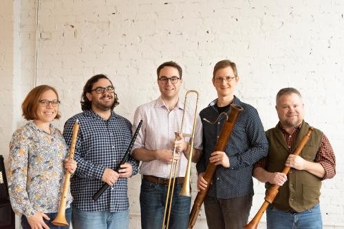 A quintet of musicians pose in a studio with white brick walls. They hold various wind instruments, some of which appear to be historical or period instruments. 