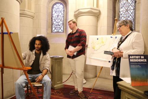 A man with long frizzy hairs sits at an easel with a young man and an older woman look over his shoulder.