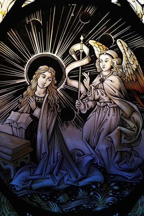 A close-up of a stained-glass window depicting the Annunciation. On the left, the Virgin Mary kneels, with a halo and her hands folded in prayer, looking down and to the side. On the right, the angel Gabriel kneels, holding a staff with a scroll unfurling from the top. Both figures are set against a dark background with light radiating from behind Mary. The bottom of the image shows a field of green plants.
