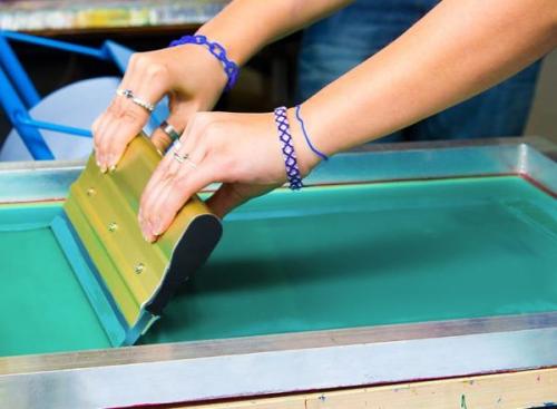 close-up shot of a person's hands holding a gold-colored squeegee over a screen printing frame. The screen is covered with a layer of teal-colored ink. The person's forearms are visible, and they are wearing a few rings and two blue woven bracelets.